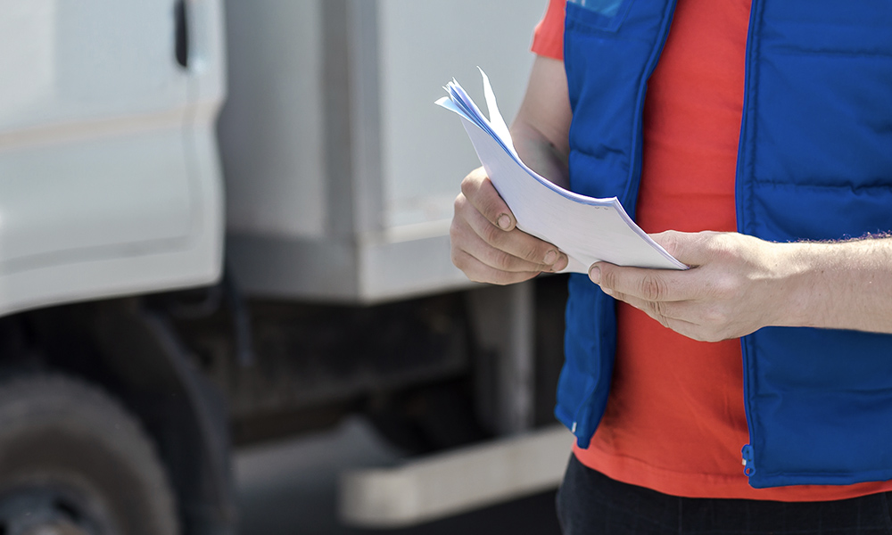 Blog - Driver in Uniform Making Notes in Document and a Delivery White Truck is Parked Behind Him
